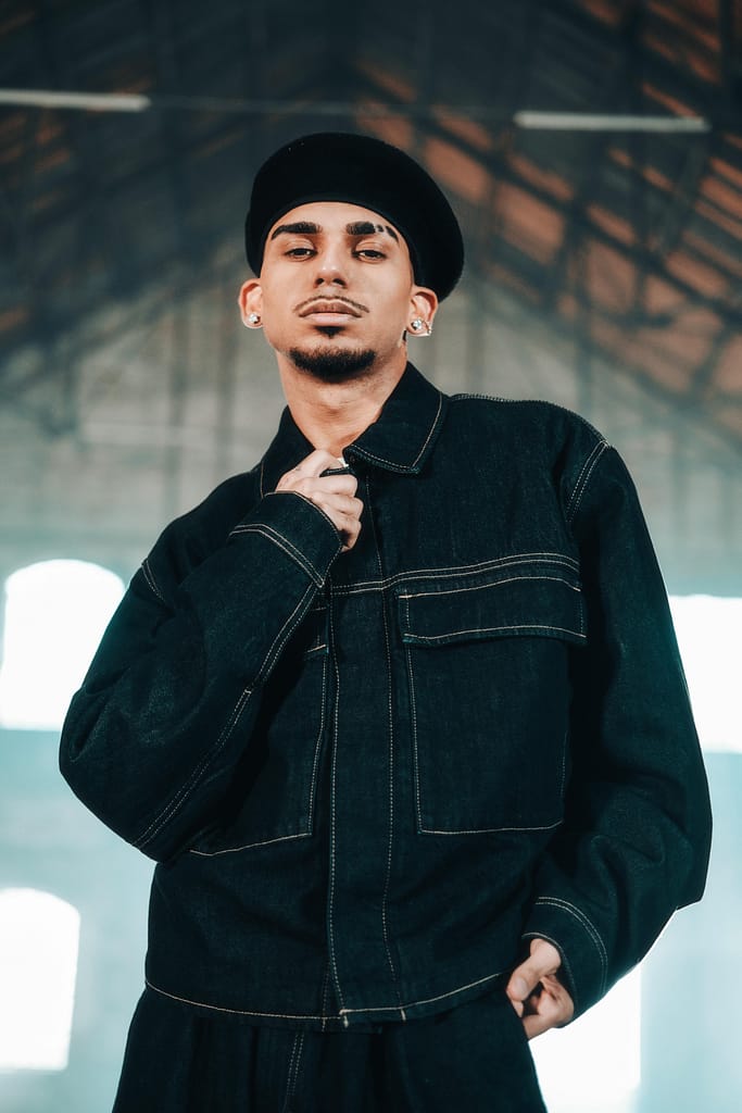 Young man wearing a denim jacket and hat posing confidently indoors, captured in a modern fashion portrait.