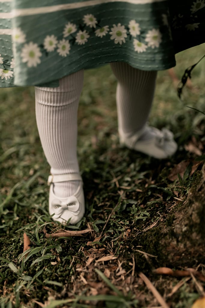 Close-up of a child's legs in white shoes and a floral dress standing on grassy ground.