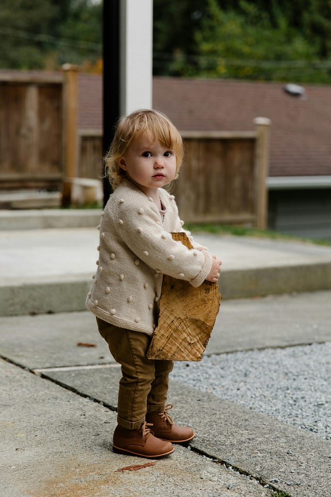 A cute toddler in knitwear holding a wooden log outdoors with a curious expression.