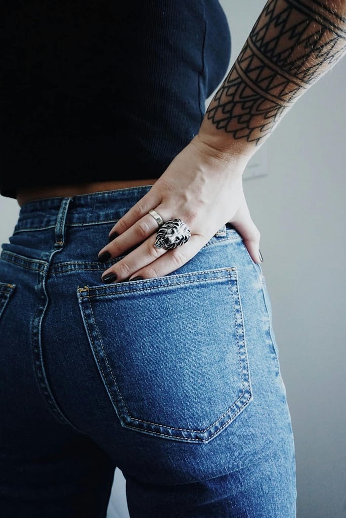 Close-up of a woman's hand with rings on her tattooed arm wearing jeans.