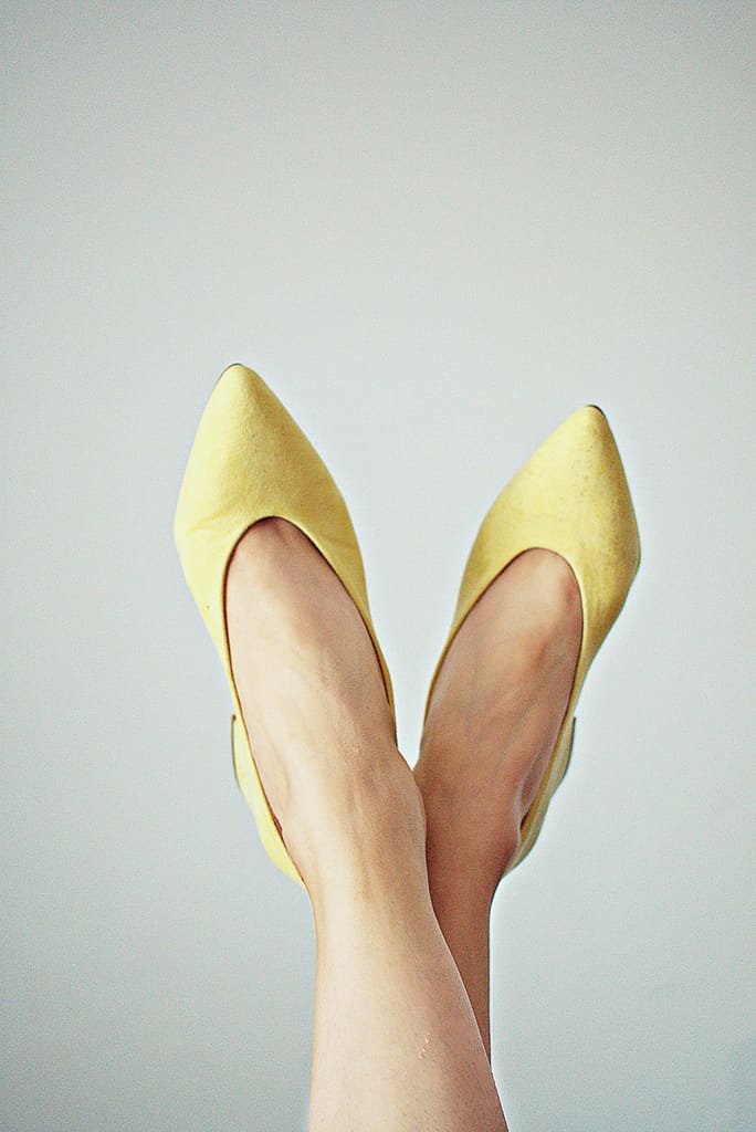 Elegant yellow pointed shoes worn by a woman with legs crossed against a plain background.